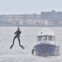 An NYPD Scuba Team member dangles above the water with an NYPD patrol boat in the background.
