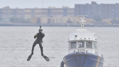 An NYPD Scuba Team member dangles above the water with an NYPD patrol boat in the background.