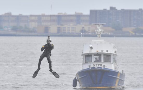 An NYPD Scuba Team member dangles above the water with an NYPD patrol boat in the background.