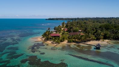 An aerial view of Bocas del Toro in Panama showing clear water and reefs leading up to a sandy beach with a few buildings at the waters' edge