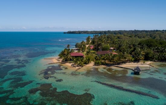 An aerial view of Bocas del Toro in Panama showing clear water and reefs leading up to a sandy beach with a few buildings at the waters' edge