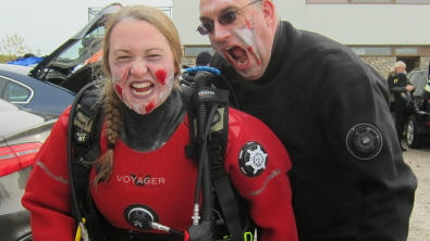 Two divers in zombie makeup, one diver pretends to bite the other