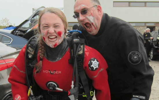Two divers in zombie makeup, one diver pretends to bite the other