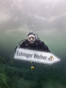 A diver underwater holding an arrow-shaped sign that says Echinger Weiher