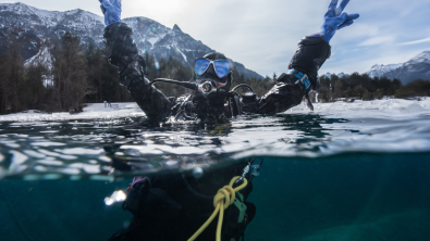 An over under shot of a diver in a dry suit raising their hands out of the water and looking joyful. The shoreline in the background is covered in snow