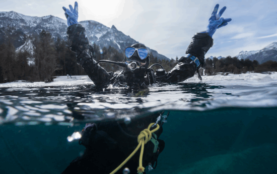 An over under shot of a diver in a dry suit raising their hands out of the water and looking joyful. The shoreline in the background is covered in snow