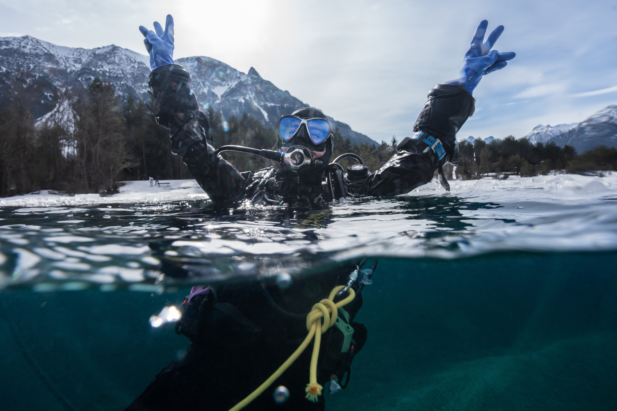An over under shot of a diver in a dry suit raising their hands out of the water and looking joyful. The shoreline in the background is covered in snow
