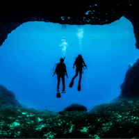 Two divers in silhouette hover in the blue framed by a large swim through in the reef