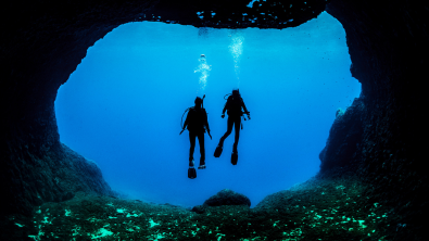 Two divers in silhouette hover in the blue framed by a large swim through in the reef