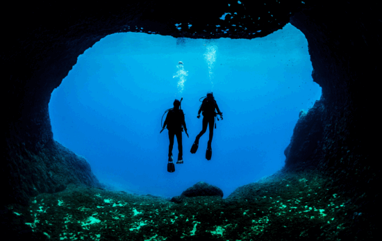 Two divers in silhouette hover in the blue framed by a large swim through in the reef