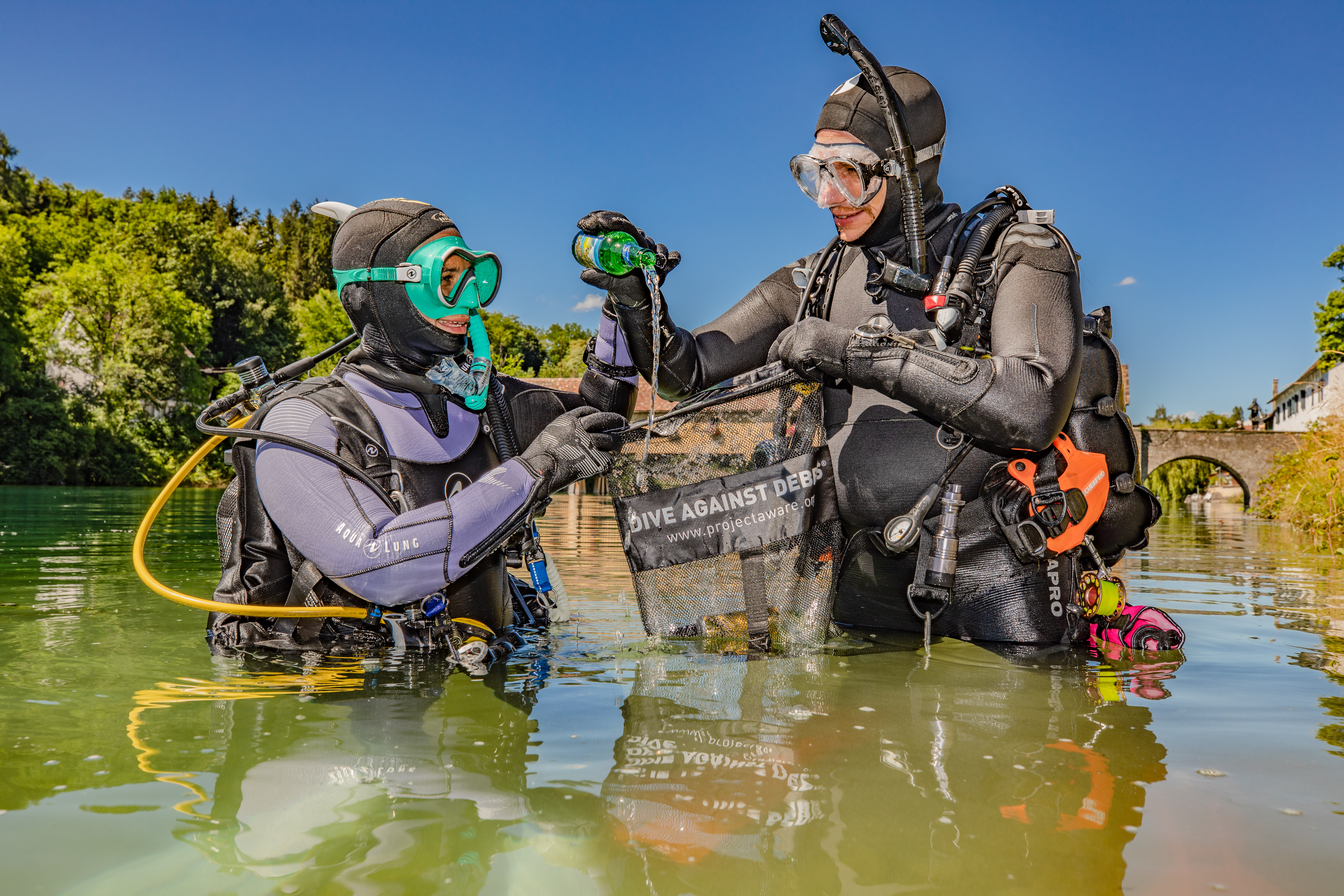 Twee duikers staan in heupdiep water en houden een Dive Against Debris tas vast