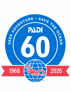 A large blue circle with the words "Seek Adventure. Save the Ocean" surround a smaller circle with a large "60" and the word "PADI" above it. Below the circle is a red banner. The PADI logo is in the middle with "1966" to the left and "2026"to the right