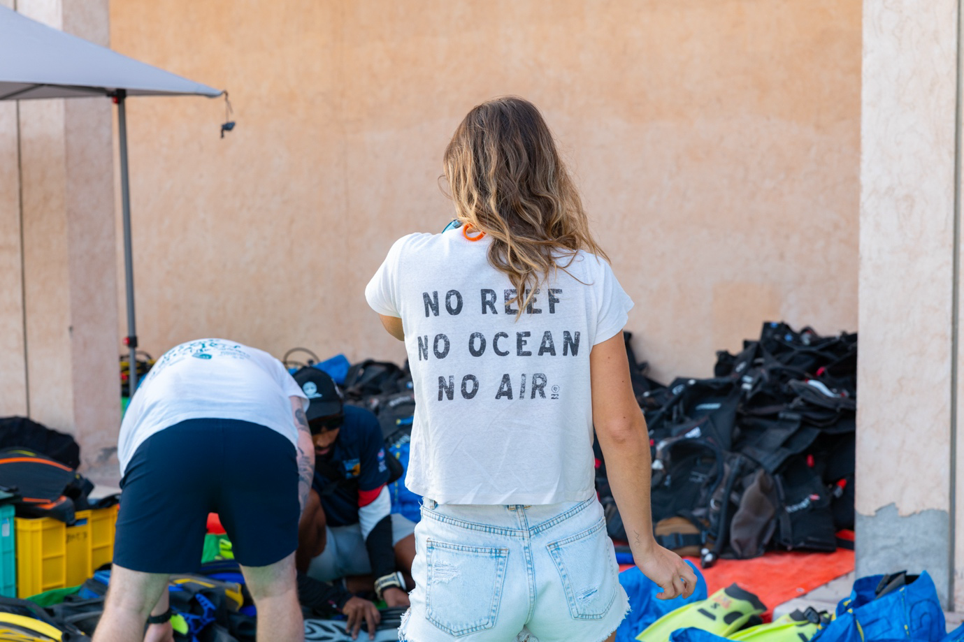 Diver standing in front of a pile of dive gear with a No Reef, No Ocean, No Air tshirt