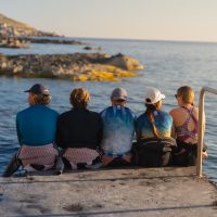 six divers sit on the end of a pier looking out into the ocean while vacationing in Malta