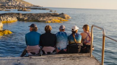 six divers sit on the end of a pier looking out into the ocean while vacationing in Malta
