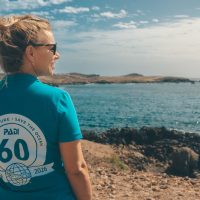 A woman wears a 60th anniversary PADI shirt and walks on a beach, looking back over her shoulder at the camera.
