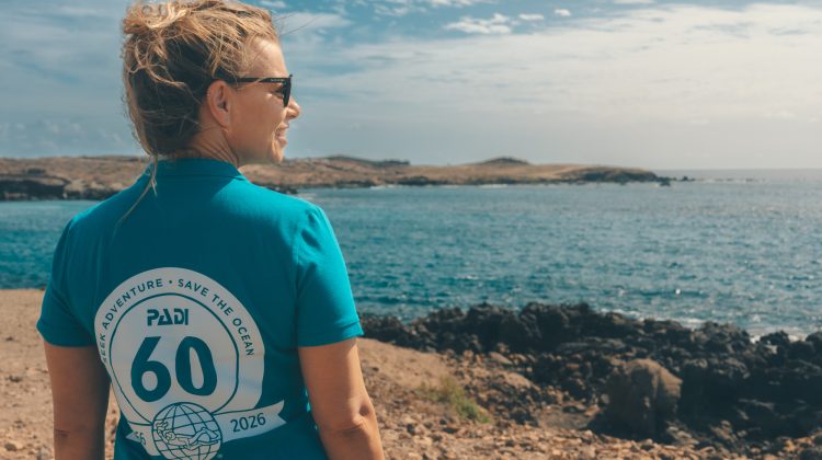 A woman wears a 60th anniversary PADI shirt and walks on a beach, looking back over her shoulder at the camera.