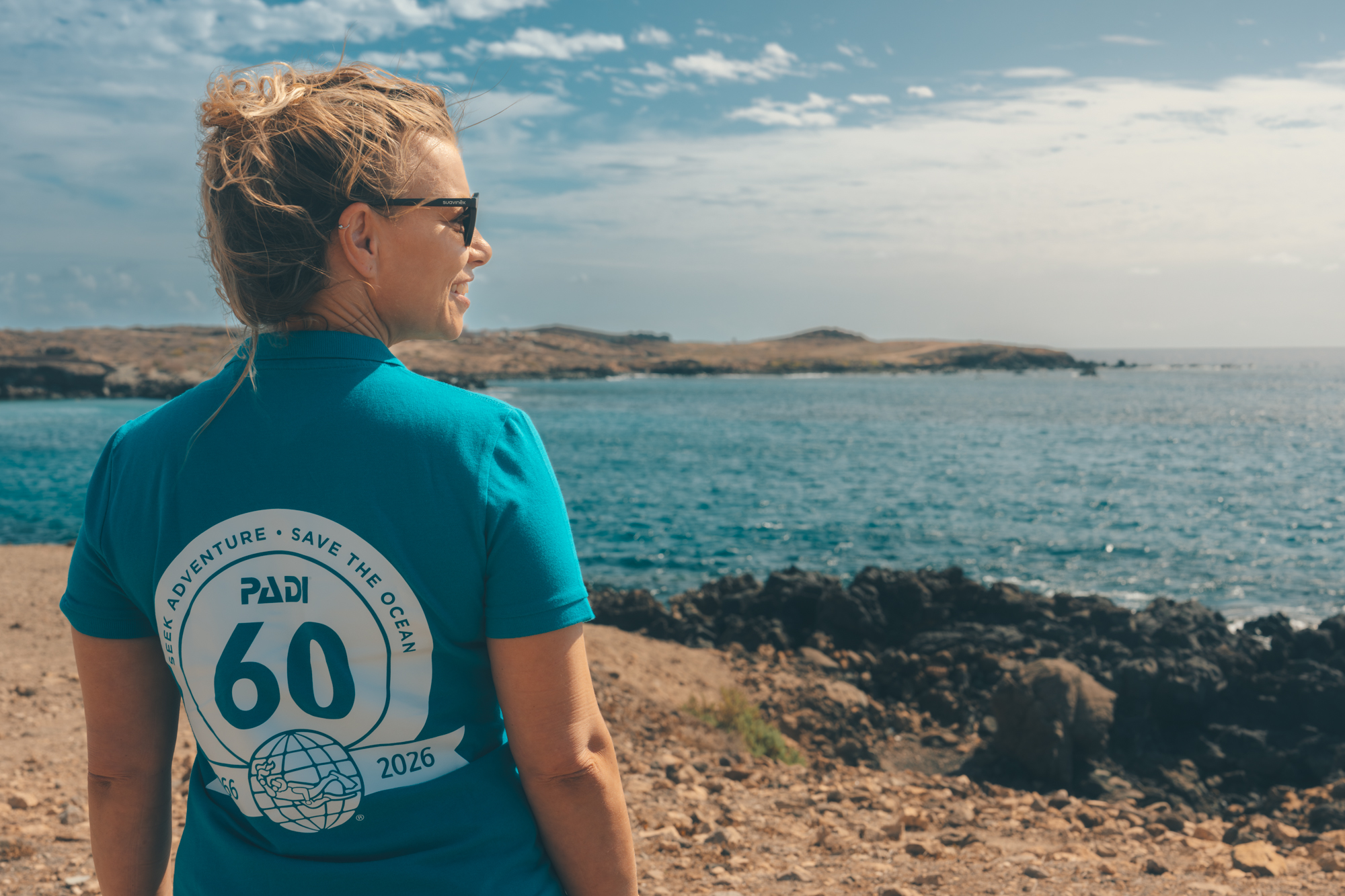 A woman wears a 60th anniversary PADI shirt and walks on a beach, looking back over her shoulder at the camera.
