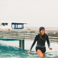 India Black walks out of the ocean wearing a rashguard and buff on her head. She is smiling.