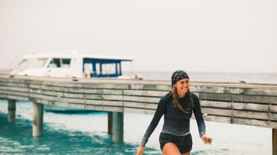 India Black walks out of the ocean wearing a rashguard and buff on her head. She is smiling.