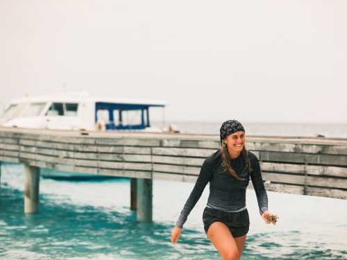 India Black walks out of the ocean wearing a rashguard and buff on her head. She is smiling.