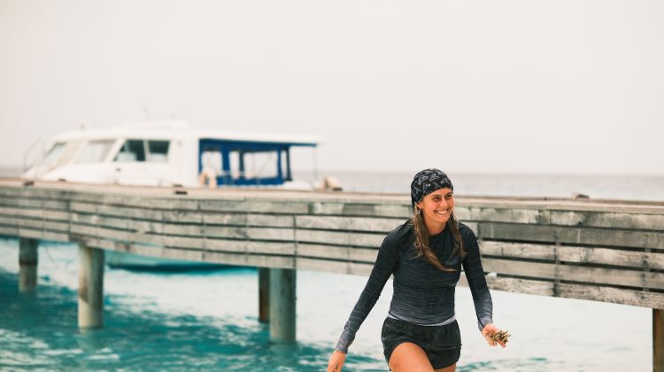 India Black walks out of the ocean wearing a rashguard and buff on her head. She is smiling.