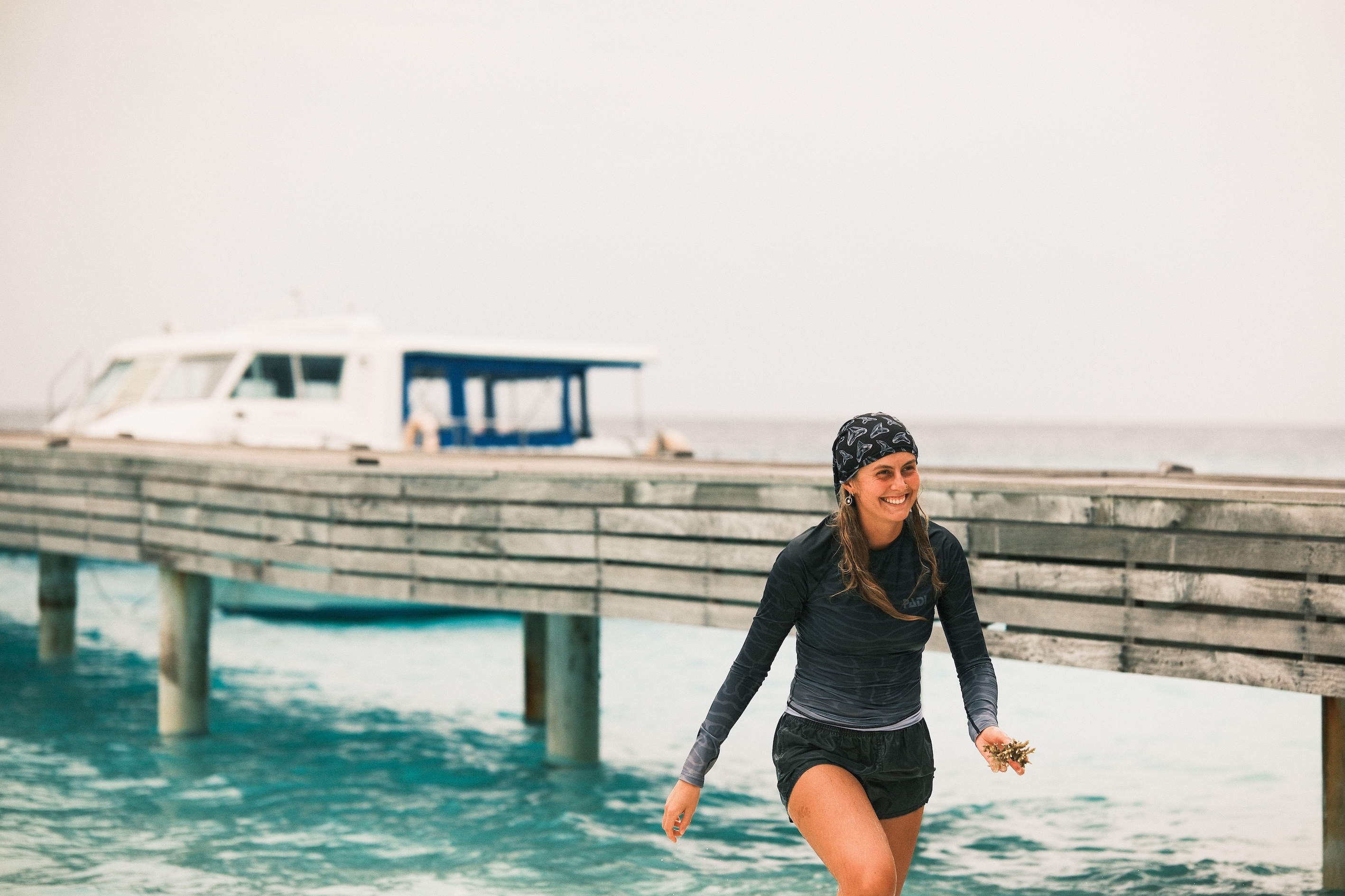 India Black walks out of the ocean wearing a rashguard and buff on her head. She is smiling.