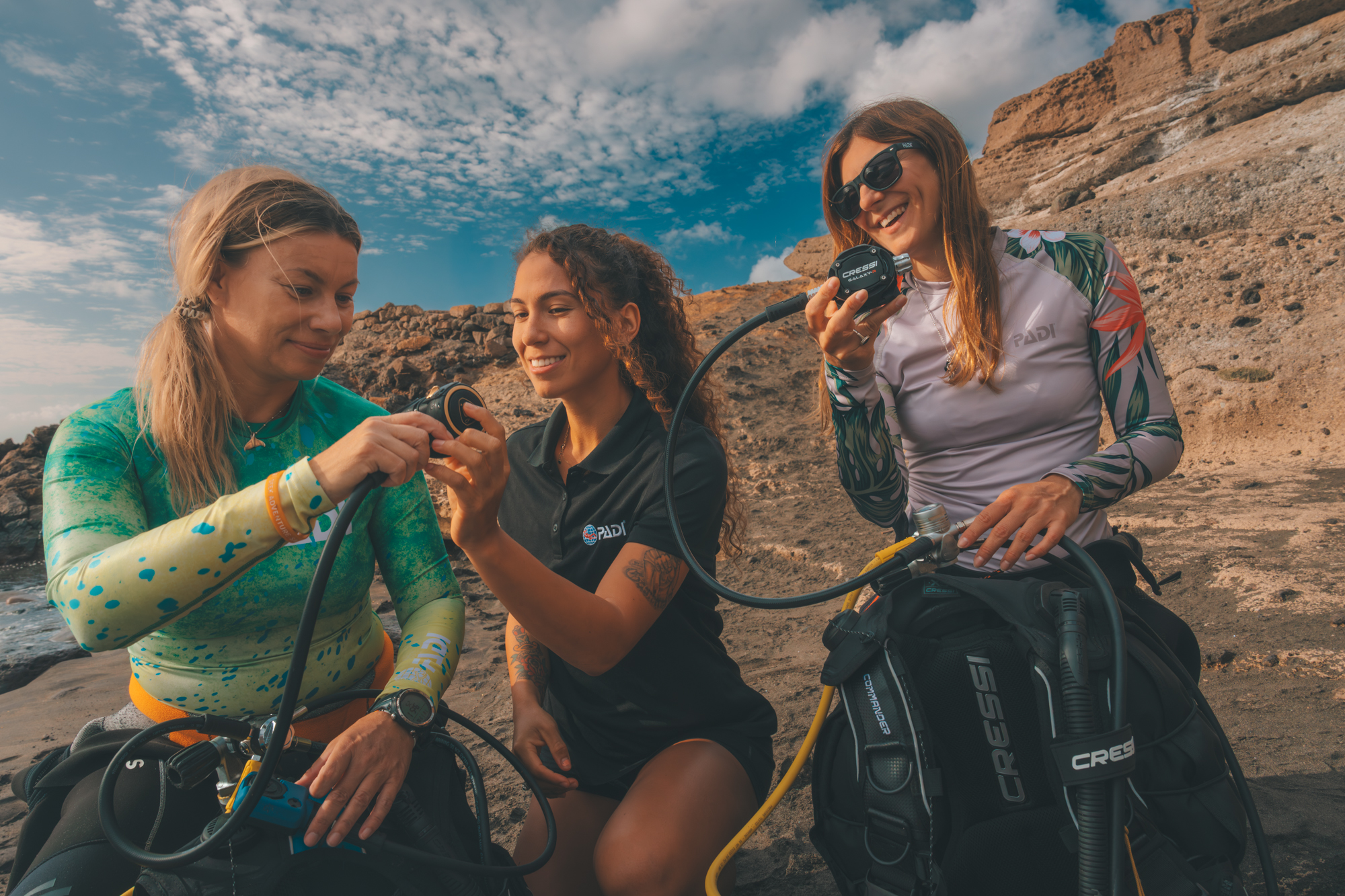 PADI Equipment Specialists Three female scuba divers on a beach learn how to inspect scuba diving gear as part of the PADI Equipment Specialist course