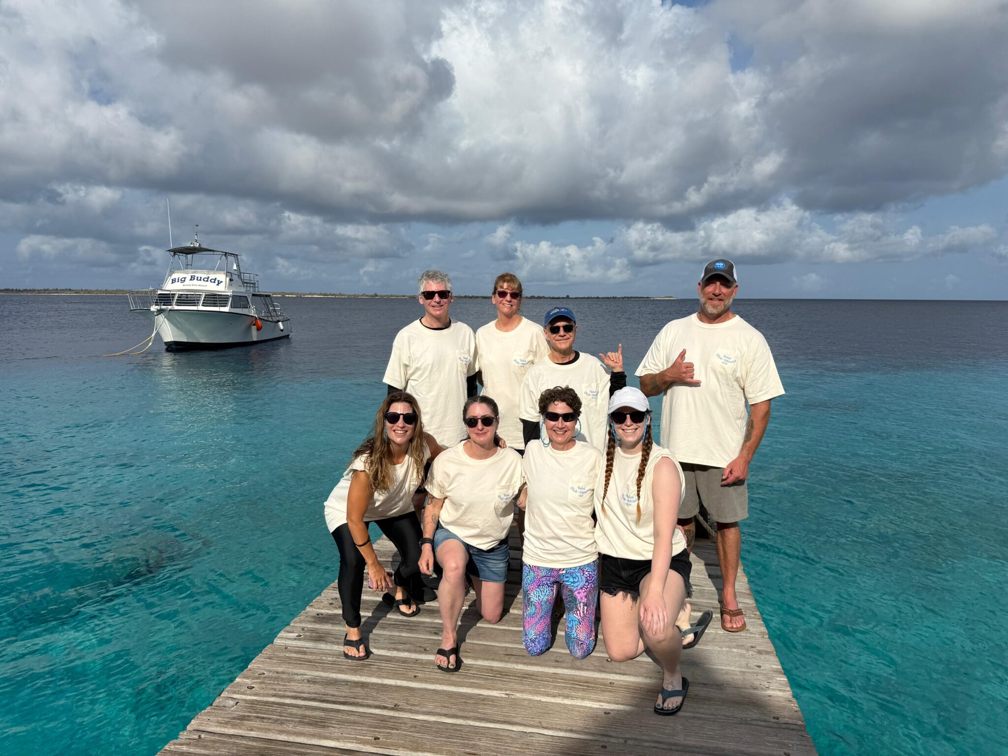 A group of smiling people wearing matching, cream-colored shirts, stand on a dock. A Buddy Dive Bonaire dive boat is in the background