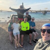 A group of divers sit on the bed of a pickup truck while another holds the camera for the selfie. A large pier is in the background