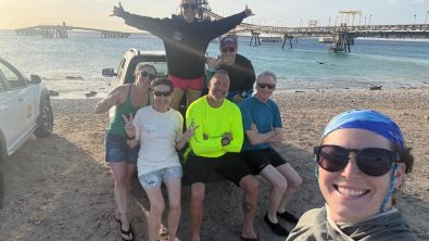 A group of divers sit on the bed of a pickup truck while another holds the camera for the selfie. A large pier is in the background