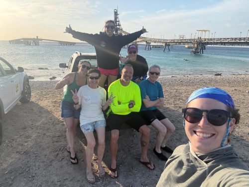 A group of divers sit on the bed of a pickup truck while another holds the camera for the selfie. A large pier is in the background