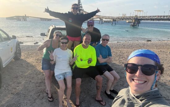 A group of divers sit on the bed of a pickup truck in Bonaire while another holds the camera for the selfie. A large pier is in the background
