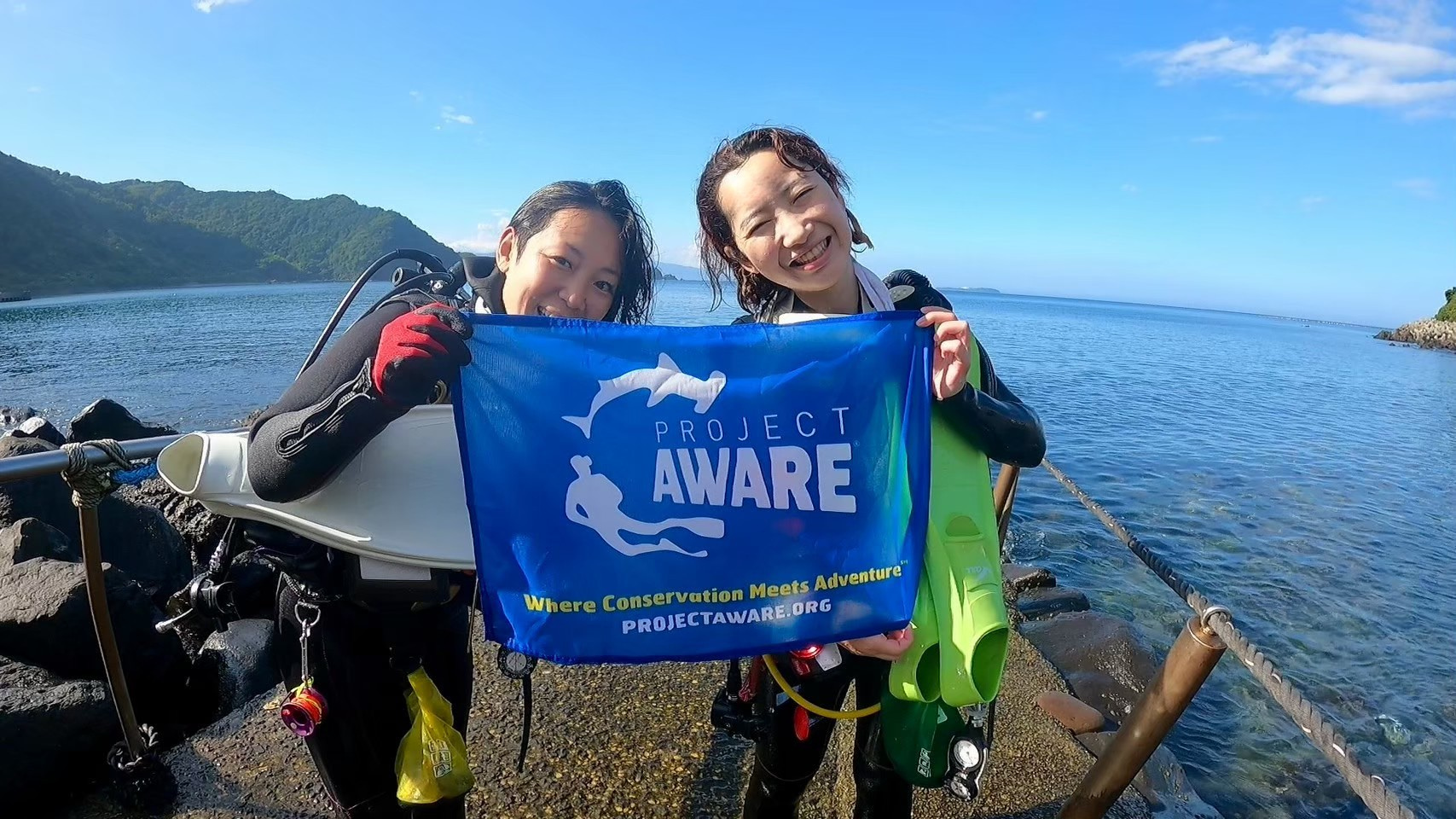 Ambassadiver & Ocean Torchbearer Award winner Yumi Shirai with another diver standing in front of the ocean holding a Project AWARE flag